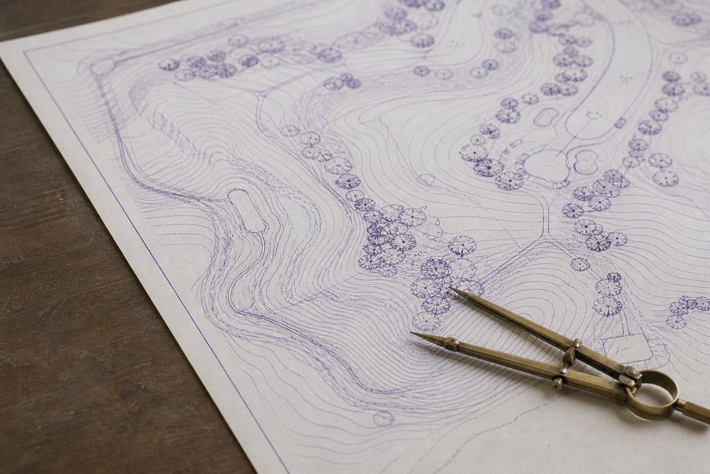 Close view of a golf course routing plan on drafting paper, hand-inked contour lines converging beneath a brass drafting compass.