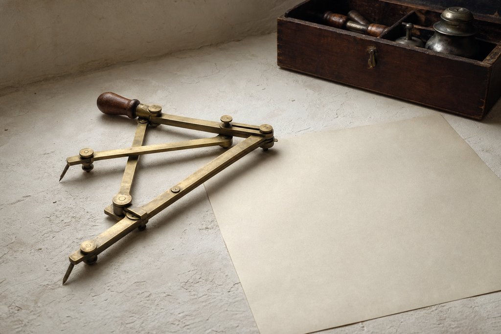 A brass pantograph copying instrument lying part-folded on a plastered surface beside a blank sheet of drafting paper.
