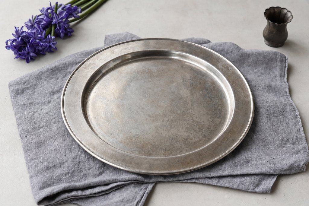 A plain unengraved silver salver with a wide rim lying flat on folded grey cloth, the surface softly tarnished.