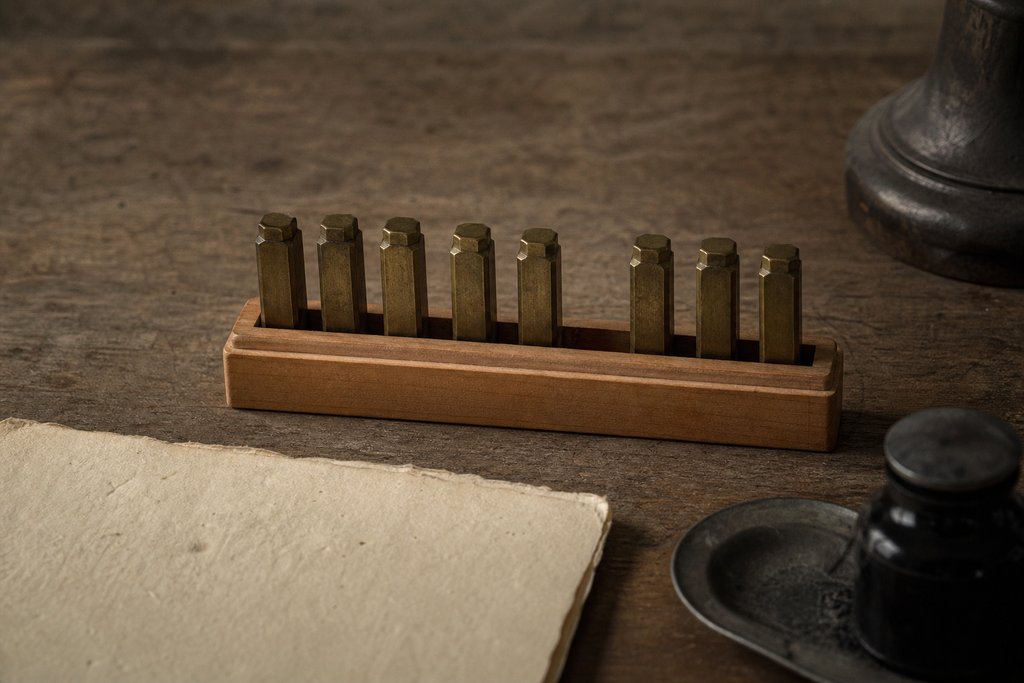 A row of small brass numbering stamps standing in a boxwood tray with one position left empty, their faces turned away.